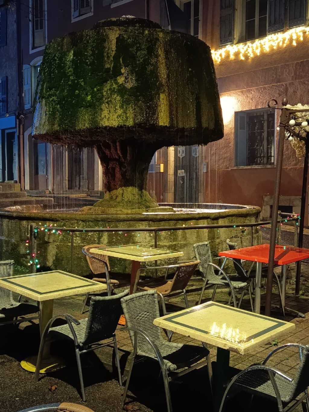 La terrasse illuminée en soirée, devant la grande fontaine moussue de Barjols